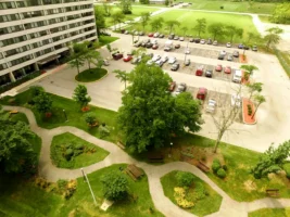 Aerial view of an apartment building, a parking lot with cars, winding paths, benches, and trees in a landscaped green area.