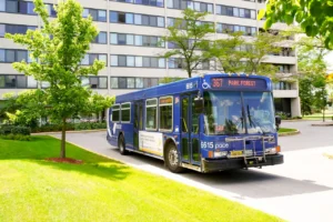 A blue city bus labeled “Pace 367 Park Forest” is parked on a road beside a modern apartment building surrounded by green trees and grass.