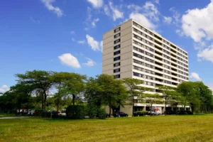 A tall, beige apartment building with many windows stands behind rows of trees and a grassy field under a blue sky with scattered clouds.