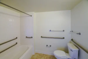 Accessible bathroom with a bathtub, toilet, and multiple wall-mounted grab bars on white walls, plus a shower curtain rod and beige tile floor.