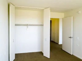 Empty closet with double white doors open, a metal hanging rod, and a shelf inside; adjacent to an open door leading to a hallway, with beige carpet and white walls.