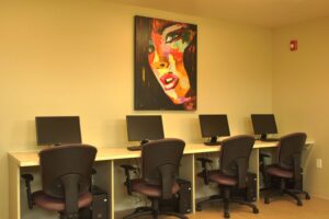 Four computer workstations with black chairs are lined up against a beige wall, beneath a colorful painting of a woman’s face.