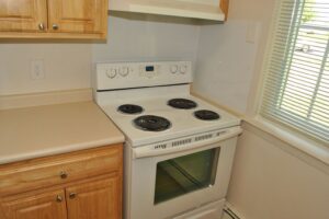 White electric stove with four coil burners and an oven, set between wood cabinets and a window with blinds in a kitchen.