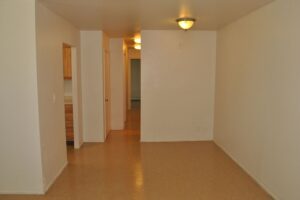 Empty room with beige floor, white walls, two ceiling lights, and three doorways; one leads to a kitchen area with wooden cabinets.