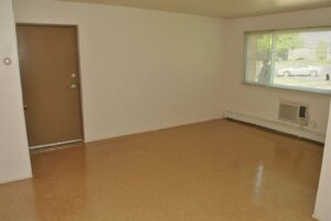 Empty room with tan linoleum flooring, white walls, a brown door, a large window with blinds, and a wall-mounted air conditioning unit beneath the window.