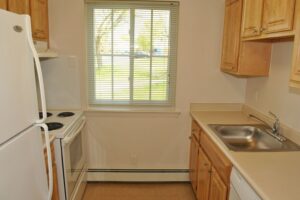Small kitchen with light wood cabinets, white appliances, electric stove, single sink, beige countertops, and a window with blinds letting in natural light.