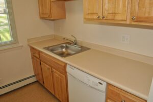 Small kitchen with light wood cabinets, beige countertop, stainless steel sink, dishwasher, and a window with blinds allowing natural light in.