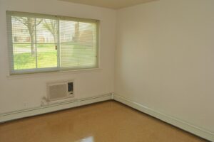 Empty room with beige linoleum floor, white walls, a window with blinds, and a wall-mounted air conditioning unit below the window.