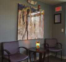 Two purple chairs and a small table with a colorful lamp sit beneath an abstract painting in a waiting room with gray walls and an exit sign above.