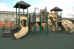 Playground with slides, climbing structures, and platforms on green safety flooring, set in front of a two-story building with a fenced yard.