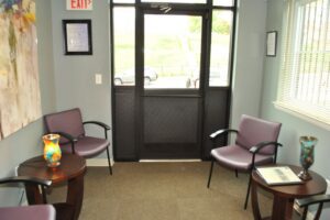 Small waiting room with four purple chairs, two side tables with decorative vases, carpeted floor, and a glass door leading outside. Sunlight comes through windows on the right.