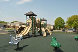 Outdoor playground with tan slides, climbing structures, and two spring riders shaped like a plane and a frog, surrounded by a rubberized surface and fences, with trees and buildings in the background.