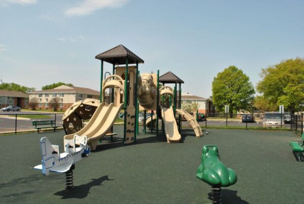 Outdoor playground with tan slides, climbing structures, and two spring riders shaped like a plane and a frog, surrounded by a rubberized surface and fences, with trees and buildings in the background.