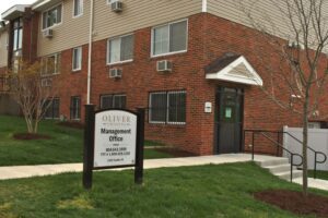 A brick apartment building with a sign in front reading “Oliver Crossing Management Office” and contact information, surrounded by grass and a sidewalk.