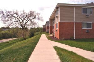 A sidewalk runs alongside a grassy area and a two-story brick and siding apartment building with several windows and air conditioning units.