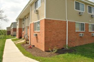 Two-story apartment building with tan siding, red brick lower walls, several window air conditioners, and a sidewalk running alongside landscaped grass.