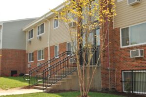 A brick and siding apartment building with several windows, a staircase with railings, air conditioning units, and a small tree in front.