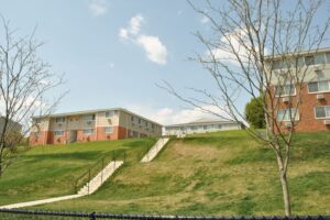 Apartment buildings with brick and siding exteriors sit on a grassy hill, separated by a concrete stairway. Leafless trees are in the foreground under a partly cloudy sky.