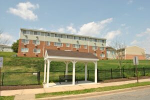 Covered bus stop with bench on a sidewalk in front of a fenced grassy hill and a three-story apartment building on a sunny day.