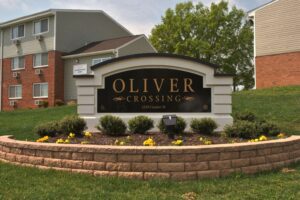 A black and gold sign reading “Oliver Crossing, 1329 Coulter St” stands in front of apartment buildings, surrounded by flowers and shrubs.