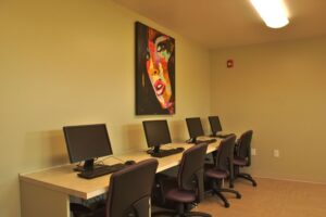 Four desktop computers with keyboards and mice are arranged on a counter in a small room. A colorful painting of a face hangs on the wall above the stations.