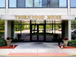 Entrance of a building with large glass doors and “THORNWOOD HOUSE” in gold letters above. Two potted plants flank the entrance. Reflections of cars and trees are visible in the glass.