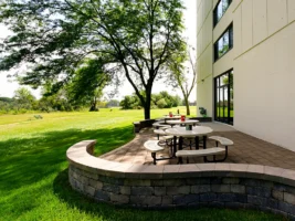 Round outdoor tables with benches are set on a paved patio beside a modern building, bordered by a low stone wall and surrounded by green grass and trees.