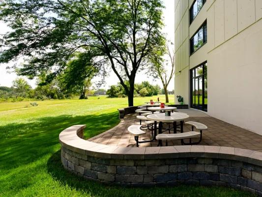 Round outdoor tables with benches are set on a paved patio beside a modern building, bordered by a low stone wall and surrounded by green grass and trees.