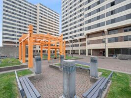 A courtyard with benches, a wooden pergola, and two tall apartment buildings in the background under a blue sky.