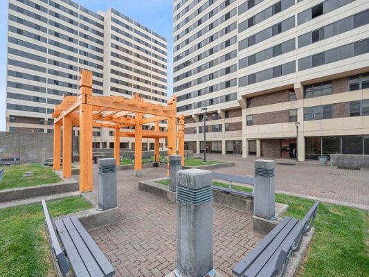 A courtyard with benches, a wooden pergola, and two tall apartment buildings in the background under a blue sky.