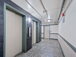 Two closed elevator doors in a brightly lit hallway with patterned flooring, wall-mounted lights, and a white door at the end of the corridor.