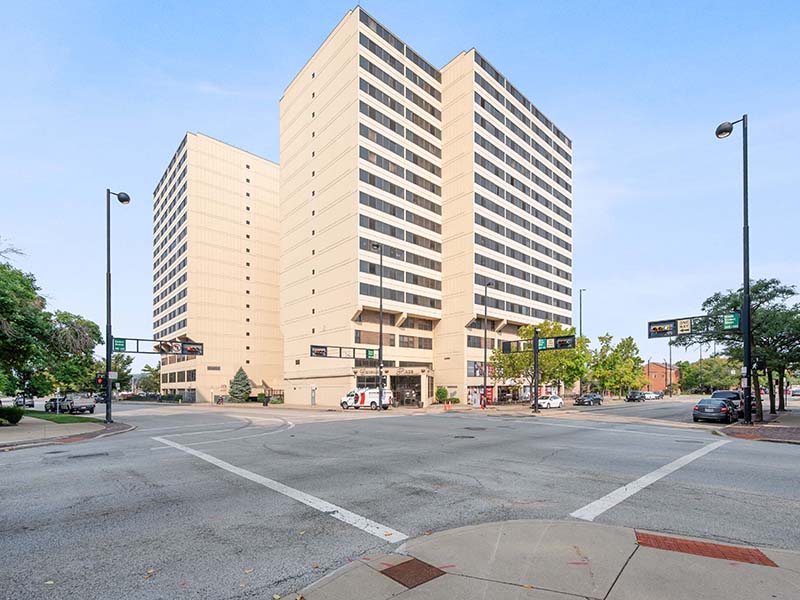 A tall, beige office building with multiple floors stands at the corner of a city intersection on a clear day.