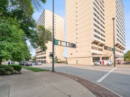 Two tall beige buildings stand along a city street with a sidewalk, traffic lights, cars, and a white van visible in the scene. Trees line the left side of the image.