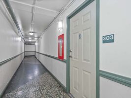 A hallway with white walls, green trim, a door labeled 303, a fire hose cabinet, and overhead lights.