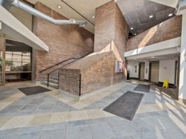 Modern building lobby with tile floors, brick walls, large windows, overhead vents, and carpeted mats. An elevator, bulletin board, and caution sign are visible. No people present.