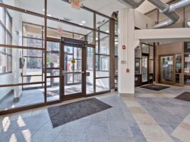 Lobby entrance with glass doors, tiled floor, black mats, and visible ceiling ducts. Sunlight streams in from large windows; an exit sign hangs above the main door.