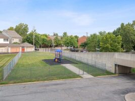 Fenced playground with a blue and yellow play structure on mulch, surrounded by grass, beside a concrete ramp and adjacent to a street with trees and houses in the background.