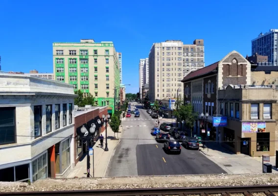 City street view from an elevated position, showing buildings, parked cars, and trees under a clear blue sky.
