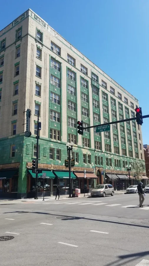 A large beige and green building with storefronts at street level and cars and pedestrians at a city intersection under a clear blue sky.