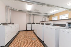 A commercial laundry room with multiple white washing machines and dryers aligned against the walls, featuring a patterned tile floor and exposed ceiling pipes.