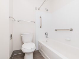 A bathroom featuring a white toilet next to a bathtub with installed grab bars, a showerhead, and a towel rack.