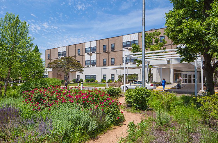 Exterior view of a multi-story brick building with a covered entrance, surrounded by trees, a garden of red roses, and a dirt walking path.
