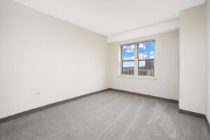 Empty room with plain walls, grey carpet, and a window overlooking city buildings.