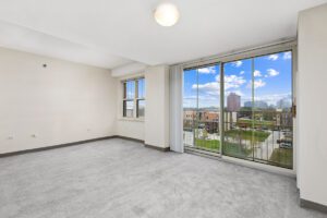 Empty carpeted room with a single window and a large sliding glass door leading to a balcony overlooking a city street.