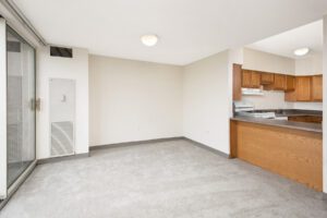 Empty room with gray carpet and white walls, featuring a sliding glass door to a balcony and an open kitchen with wood cabinets and a breakfast bar.