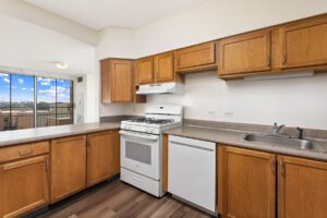 A kitchen with light wood cabinets, white appliances, and a stainless steel sink, viewed from an open living area with a balcony door.