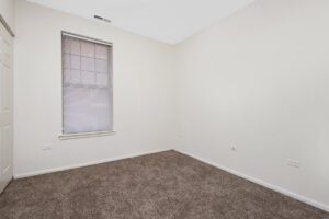 Empty corner of a room with beige walls, brown carpet, and a single window with closed blinds.