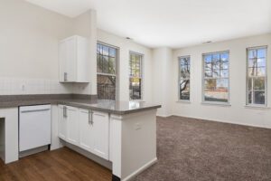 A kitchen with white cabinets, speckled countertops, and a white dishwasher, opening into a carpeted living area with four windows.