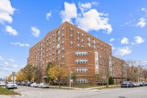 A large, multi-story brick apartment building on a corner street intersection with parked cars along the curb.