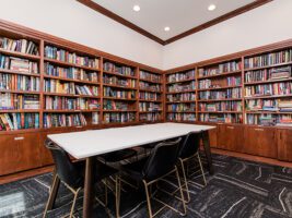 A white rectangular table surrounded by four black chairs in a library with floor-to-ceiling wooden bookshelves on two walls.